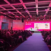 Auf dem Bild ist eine große Bühne mit Mittelgang zu sehen. Links und rechts davon sitzen die Gäste. Der LED Bildschirm auf der Bühne ist in pinker Farbe.