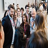 Gruppe von Menschen mit Namensschildern in einem hellen Innenraum, einige im Gespräch, andere im Hintergrund stehend