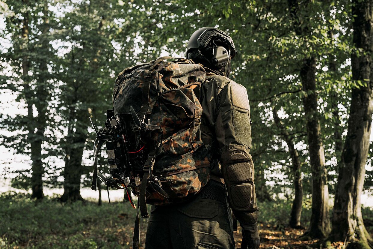 Person in Militär-Schutzkleidung transportiert eine Drohne, die am Rucksack festgemacht ist