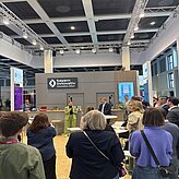 Auf dem Bild ist die Rückansicht einer Gruppe von Menschen zu sehen, welche sich stehen die Ansprache einer in grün gekleideten Frau anhören.