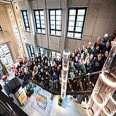 Das Bild zeigt eine große Gruppe von Menschen, die auf einer Treppe in einem modernen, offenen Gebäude stehen. Die Architektur des Gebäudes ist offen und hell, mit hohen Decken und großen Fenstern, die viel Tageslicht hereinlassen. Die Menschen scheinen ein Team oder eine Gruppe von Kollegen zu sein, die sich für ein Foto versammelt haben. Die Stimmung ist positiv und kollegial. Die Komposition des Bildes ist von oben aufgenommen, was den Eindruck von einer großen, zusammenhängenden Gruppe vermittelt.