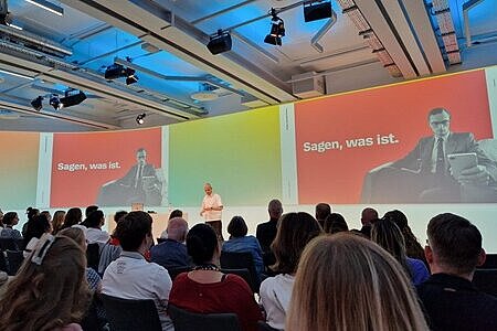 In einem voll bestuhlten Saal ist eine Bühne zu sehen, auf der Fabian Grischkat einen Vortrag hält. Er ist nur dezent aus der Ferne zu erkennen. 