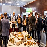 das Bild zeigt eine Gruppe von Personen bei einer Konferenz oder Netzwerkveranstaltung. Auf mehreren Tischen sind verschiedene Speisen angerichtet, und die Anwesenden stehen in Gruppen beisammen und unterhalten sich.