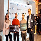 "Vier Personen stehen vor einem Hintergrund mit verschiedenen Logos. Zwei der Personen halten große Schecks in den Händen – einer mit dem Betrag '€ 750' und der andere mit '€ 500'. Die Schecks enthalten außerdem Text und Logos. Der Hintergrund ist gut beleuchtet und zeigt mehrere Markenlogos."