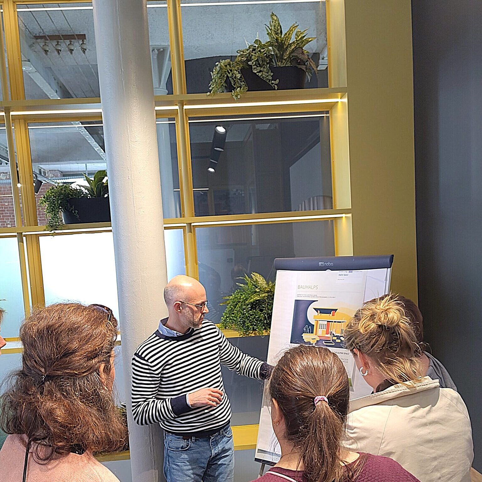 Ein Mann steht vor einem Schreibboard und erzählt etwas. Um ihn herum stehen drei Frauen. 