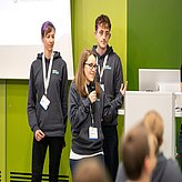 Auf dem Bild sind im Hintergrund zwei Herren in grauen Hoodies zu sehen. Im Vordergrund ist eine Dame mit Mikrofon zu sehen, welche einen Vortrag hält.