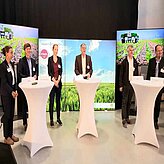 Auf einer Bühne mit Stehtischen stehen mehrere Referenten im Panelgespräch