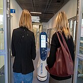 Zwei Frauen von hinten gehen in einem Flur auf einen weißen Serviceroboter mit Display und der Aufschrift »TEDIRO« zu