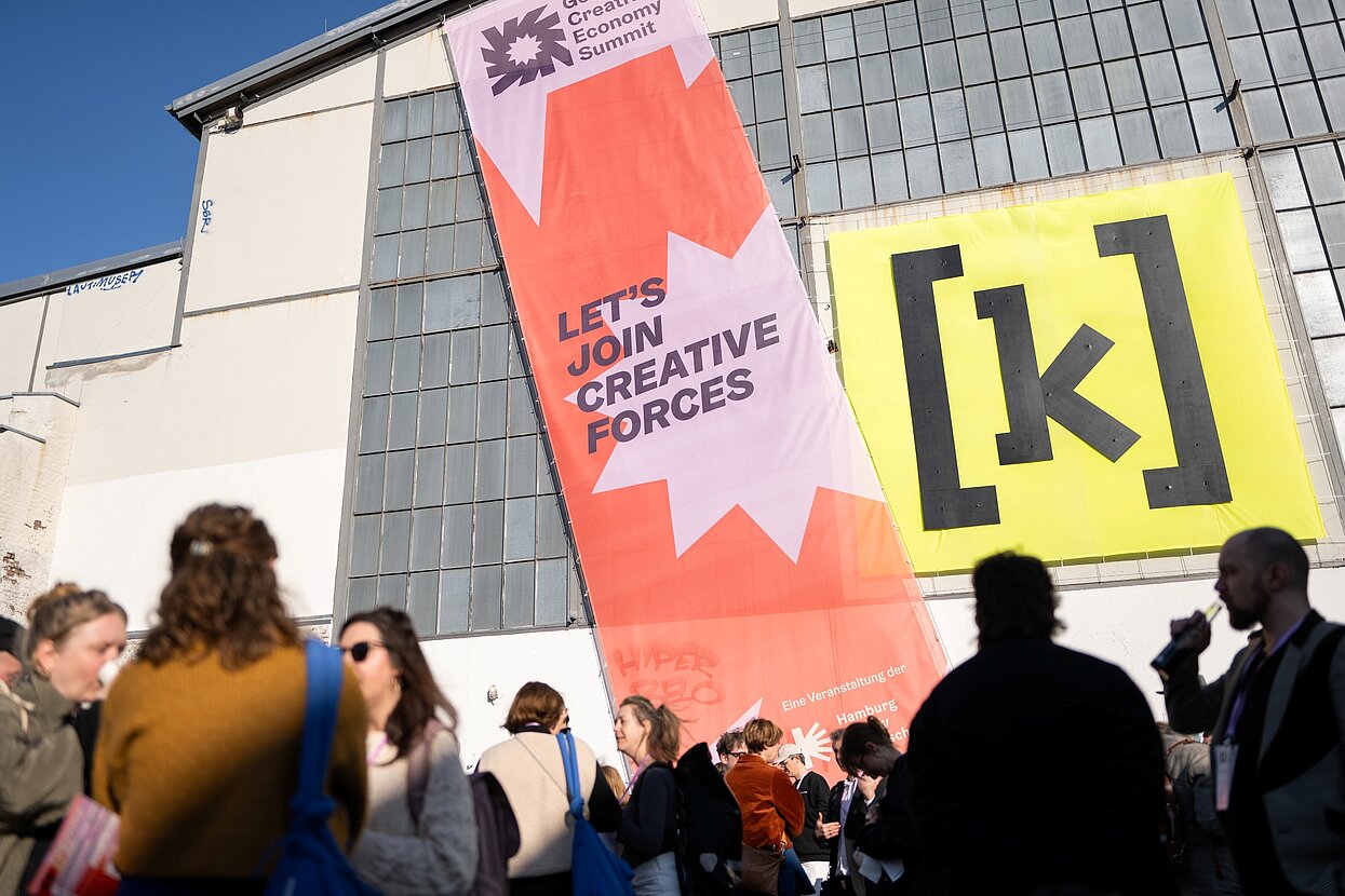 Riesiges Banner an einem Haus mit der Aufschrift "Let#s join creative forces", davor Menschen die sich unterhalten