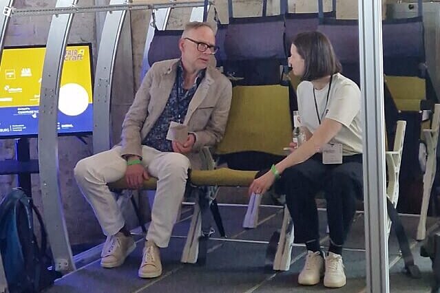 Zu sehen sind zwei Personen, die sich angeregt unterhalten. Sie sitzen auf einer Wagon-ähnlichen Bank, die einen Teil der interaktiven Ausstellung darstellt.