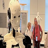 Führung: Robotik und Design im Deutschen Museum: Im Vordergrund ist der Kopf eines Ro-boters zu sehen, während dahinter ein paar Menschen stehen.