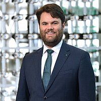Person in einem dunklen Business‑Anzug mit weißem Hemd und gemusterter Krawatte steht vor einem modernen Hintergrund aus glänzenden Glas- und Metallelementen.