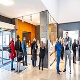 Mehrere Personen befinden sich in einem modernen, hell beleuchteten Empfangs- oder Lobbybereich. Hinter einem Tresen stehen Mitarbeitende, während einige Personen in einer Warteschlange stehen und andere sich unterhalten.