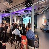 Auf diesem Bild ist eine Gruppe von Menschen zu sehen, die in einem Raum sitzen und einer Präsentation folgen. Der Raum hat moderne Beleuchtung mit violetten Akzenten und eine industrielle Decke. Vorne steht ein Präsentator in der Nähe eines Bildschirms, auf dem Folien angezeigt werden. Das Publikum scheint aufmerksam der Präsentation zu folgen.