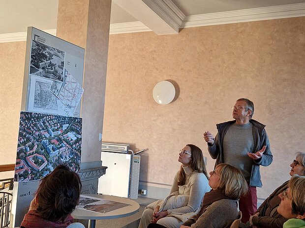 Ein Herr erklärt vor einem Publikum eine Stadtansicht. 