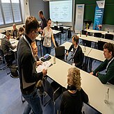 Mehrere Personen sitzen und stehen in einem Seminarraum mit Tischen, Stühlen, Flipchart und Bannern im Hintergrund