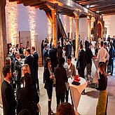 "Eine große Gruppe von Personen befindet sich bei einer Veranstaltung in Innenräumen. Der Raum ist durch hohe Holzbalken und warmes Licht geprägt. Mehrere Stehtische mit weißen Tischdecken sind im Raum verteilt, und die Anwesenden unterhalten sich in kleinen Gruppen."