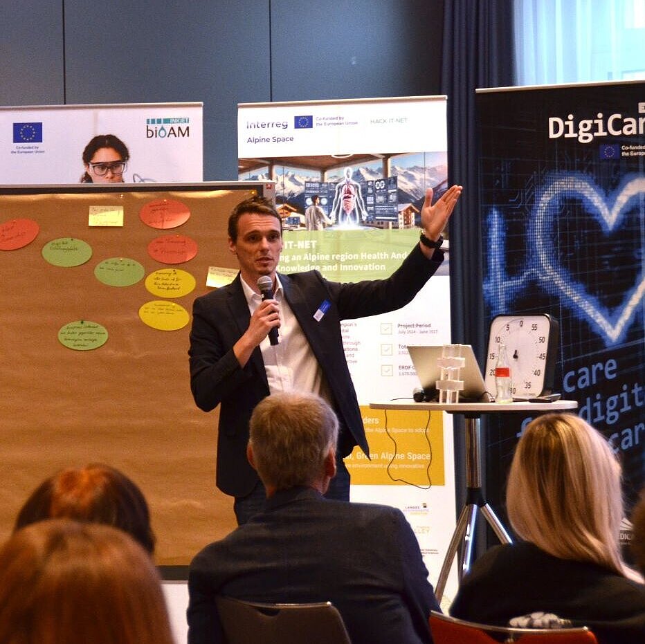 Person mit Mikrofon vor Publikum gestikuliert neben zwei Pinnwänden mit bunten Notizzetteln in einem Konferenzraum