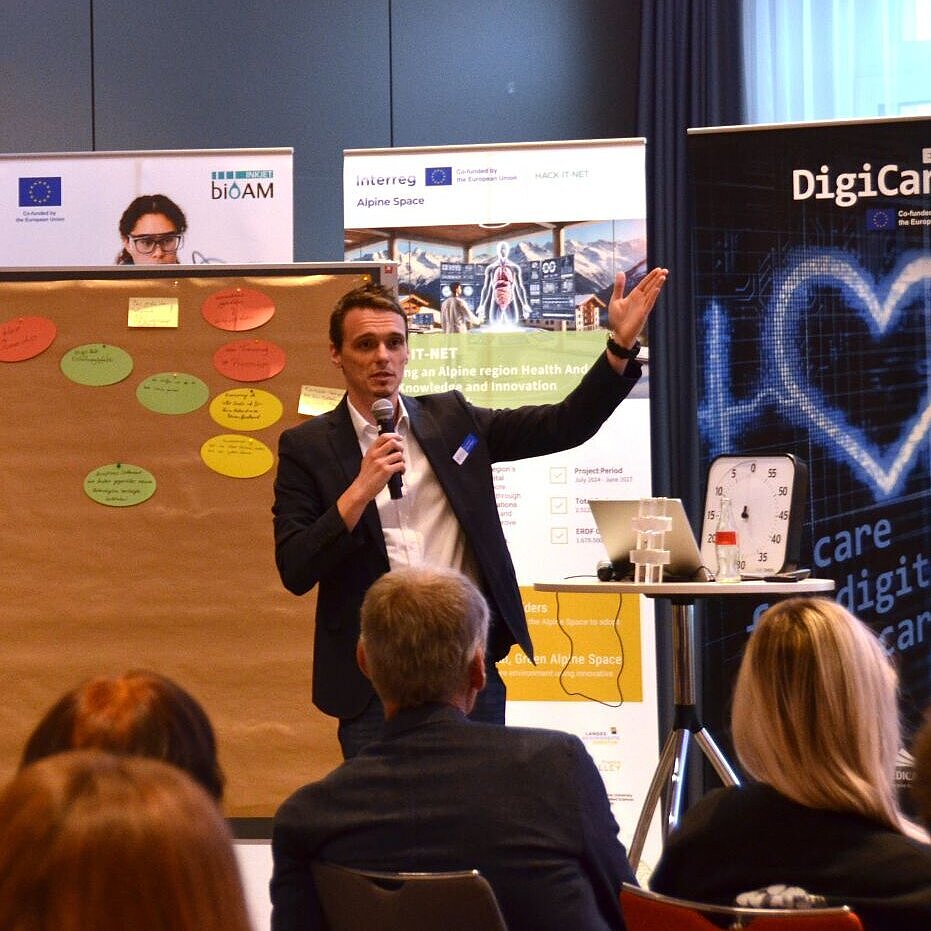 Person mit Mikrofon vor Publikum gestikuliert neben zwei Pinnwänden mit bunten Notizzetteln in einem Konferenzraum