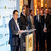 ein Herr hält eine Rede an einem Rednerpult, im Hintergund eine Logowand