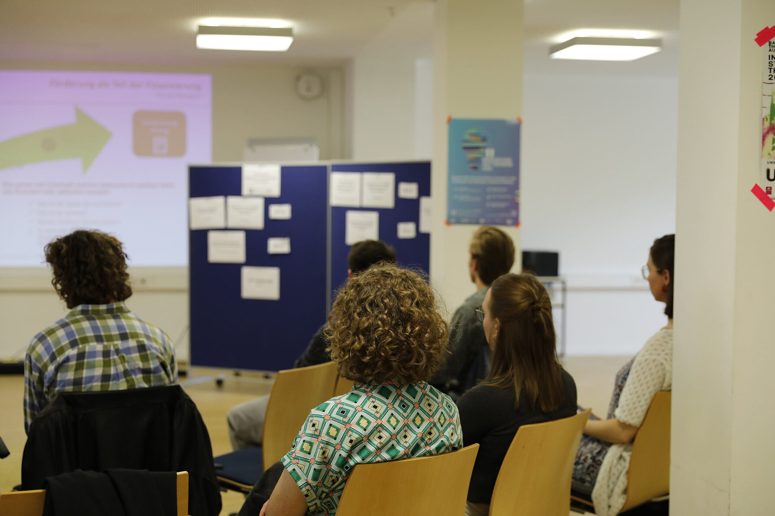 Zu sehen sind mehrere Personen von hinten. Die Personen sitzen in einem Raum auf Holzstühlen und hören einem Vortrag zu. Im Hintergrund ist eine an die Wand geworfene Folie zu erkennen.