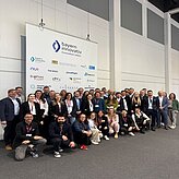 Auf dem Bild ist eine große Gruppe von Menschen zu sehen, welcher sich für ein Bild vor der Pressewand mit verschiedenen Logos positioniert haben.