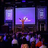 Eine Tänzerin steht mit ausgestreckten Armen auf einer Bühne und führt vor dem Publikum eine Performance auf. Hinter ihr ist eine großflächige Projektion und beleuchtete „TECH DAYS“-Logos zu sehen.