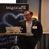 Mann in Anzug steht an Stehtisch mit Laptop und Hashtag-Symbol vor Banner mit Schriftzug 'EDIH DigiCare' und Herzgrafik
