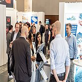 das Bild zeigt eine Gruppe von Personen auf einer Messe oder Ausstellung. Mehrere Stände mit Displays und Postern sind zu sehen, und die Anwesenden befinden sich im Gespräch.