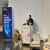 Prof. Daniel Nusser, Professor und Studiengangsleitung für Prozess- und Produktdesign, DIP-LOMA Hochschule, ist zu sehen, wie er am Pult ins Mikrofon spricht, während vor ihm eine Vase mit Blumen und ein Roll-up von Bayern Innovativ zu sehen sind.