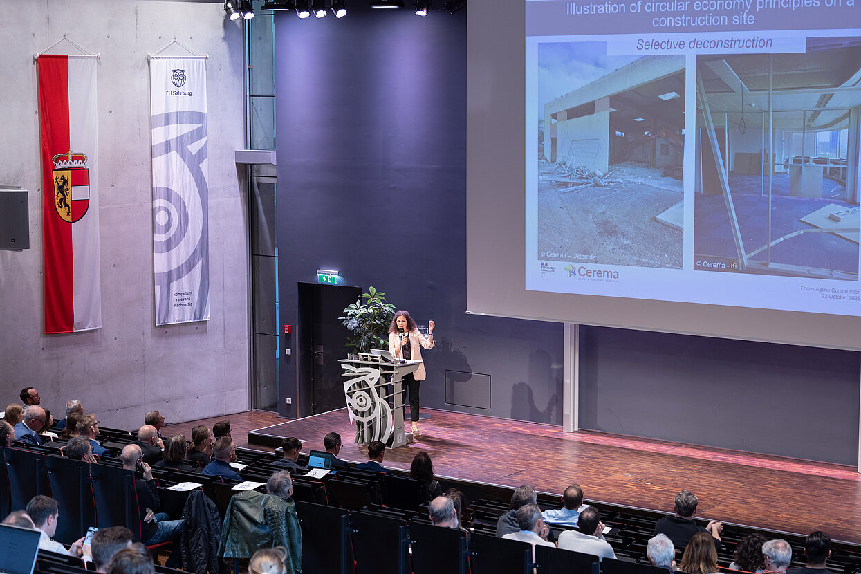 Podium im Hörsaal und Referentin, hinter ihr eine Präsentation auf der Leinwand. 
