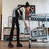 Person mit einem Exoskelett an den Beinen greift nach einer Kiste mit der Aufschrift '10' in einem Raum mit Regalen und medizinischen Postern an der Wand