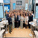 Große Gruppe von Menschen in einem modernen Messestand mit dem Logo von bayern innovativ an der Wand