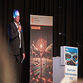 Das Bild zeigt einen Sprecher mit Mikrofon auf der Bühne des Bayerischen Batteriekongresses 2025.
