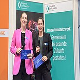 Zwei Personen stehen vor einem Hintergrund  Eine Person trägt einen pinken Blazer, die andere ein schwarzes Outfit. Beide halten eine blaue Mappe in der Hand