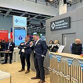 Auf dem Bild sind zwei Herren im Anzug zu sehen, welche eine Ansprache halten. Im Hintergrund ist eine bayrisch geschmückte Theke zu sehen.