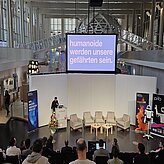 Prof. Daniel Nusser, Professor und Studiengangsleiter für Prozess- und Produktdesign, DIP-LOMA Hochschule, spricht am Pult zum Publikum, während auf dem Bildschirm der Satz „Humanoide werden unsere Gefährten sein“ zu sehen ist.