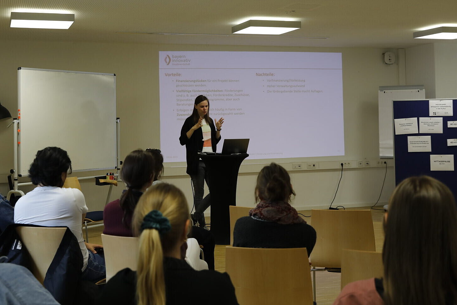 Zu sehen ist Charlotte Stegmayer vom Bereich Kreativwirtschaft der Bayern Innovativ, wie sie gerade auf deiner Bühne stehend einen Vortrag hält. Sie gestikuliert dabei mit den Händen, während das Publikum angeregt zuhört. Im Hintergrund ist eine Folie eingeblendet, auf der die Schlagworte "Vorteil" und "Nachteil" zu erkennen sind. 