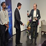 Markus Eder, Ministerialdirigent, Bayerisches Staatsministerium für Wirtschaft, Landesent-wicklung und Energie, überreicht Robert Bartl, Mitglied der Geschäftsleitung, Bayern Innova-tiv GmbH, das Mikrofon, während Christian Fayek, Leitung Projekte, bayern design GmbH, daneben steht.