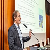 Eine Person im Anzug steht an einem Rednerpult und spricht in ein Mikrofon. Hinter der Person ist eine Präsentationsfolie auf einer Leinwand zu sehen, die Text und ein Bild enthält.