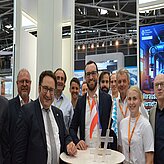 „Eine Gruppe von Personen steht um einen runden Tisch bei einer Veranstaltung in Innenräumen. Im Hintergrund sind verschiedene Ausstellungsstände und Beleuchtungselemente zu sehen, was auf eine Messe oder Konferenz hinweist.“