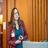 Eine Person steht vor einer Holzverkleidung und hält mehrere Gegenstände in den Händen. Die Person trägt eine weinrote Jacke, ein türkisfarbenes Oberteil und ein Namensschild. 