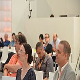 Mehrere Personen sitzen in einem Konferenz- oder Seminarraum und blicken nach vorne, vermutlich in Richtung eines Vortrags oder einer Präsentation. Der Raum ist schlicht gestaltet mit weißen Wänden und technischer Ausstattung