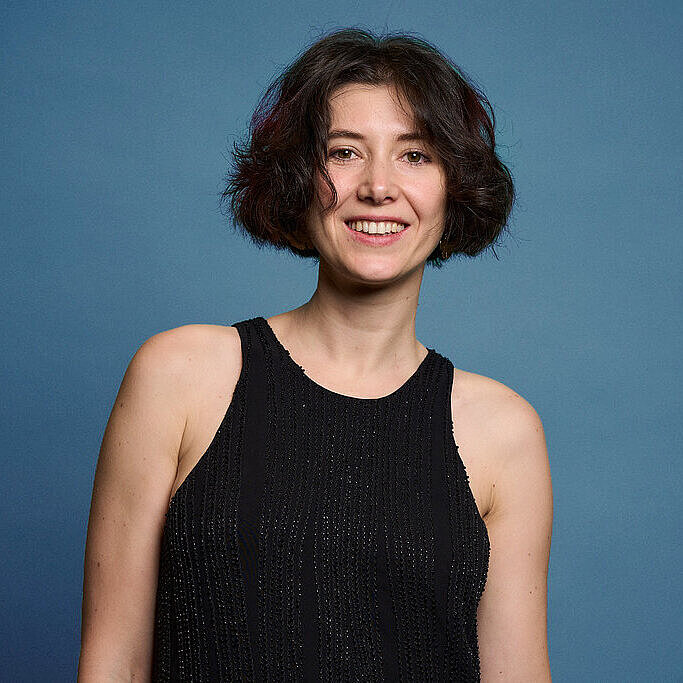 Eine Frau mit schwarzen Haaren vor einem dunkelblauen Hintergrund im einem schwarzen, glitzernen Kleid.