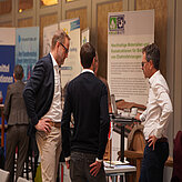Das Bild zeigt drei Männer im Gespräch an einem Messestand mit einem Poster zum Thema nachhaltige Materialien für Batterien.