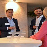 Zwei Männer in Anzügen und eine Frau in pinker Kleidung stehen an einem Stehtisch mit einem großen Hashtag-Symbol aus Holz