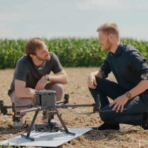 Zwei Personen knien auf einem Feld mit Mais im Hintergrund, sie sprechen miteinander. Vor ihnen ist eine Felddrohne aufgebaut.