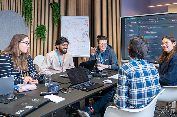 Eine Gruppe von Menschen sitzt um einen Tisch in einer Besprechung oder einer kollaborativen Arbeitssitzung. Auf dem Tisch befinden sich Laptops und Papiere, und die Personen scheinen in Diskussion vertieft zu sein. Im Hintergrund ist ein Whiteboard mit einigen Schriftzügen und Diagrammen sowie einige Pflanzen an der Wand zu sehen.