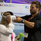 Das Bild zeigt eine Frau mit VR Brille und einen Mann der ihr hilft die Brille aufzusetzen