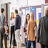 Das zweite Bild zeigt eine Gruppe von sechs Personen, die sich vor mehreren Pinnwänden oder Postern unterhalten. Die Szene spielt sich in einem Innenraum ab, vermutlich im Rahmen einer Veranstaltung wie einer Konferenz oder Ausstellung.